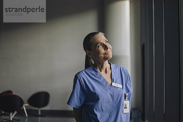 Smiling thoughtful senior female doctor in blue scrubs standing at hospital