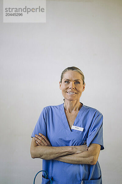 Portrait of smiling senior female doctor standing with arms crossed in front of white wall