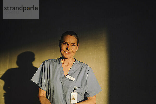 Portrait of smiling senior female doctor standing in front of wall at hospital