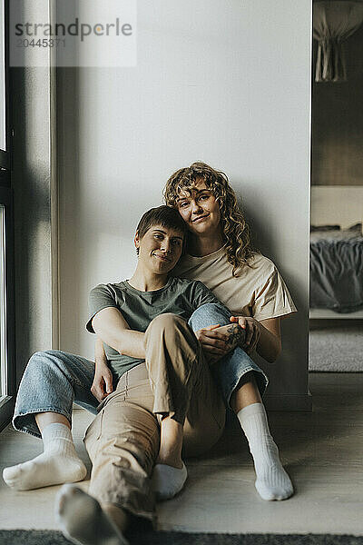 Smiling lesbian couple sitting together while leaning on wall at home