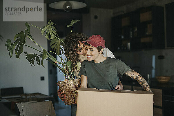 Smiling lesbian woman holding houseplant kissing girlfriend carrying cardboard box during relocation