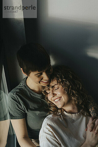 High angle view of smiling lesbian woman leaning on girlfriend while holding hands near wall at home