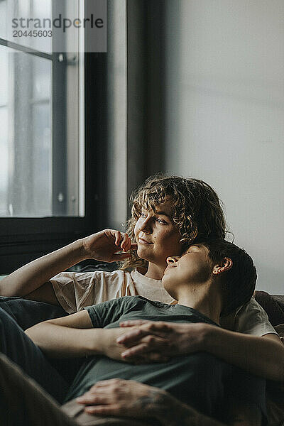 Thoughtful lesbian woman looking through window while lying down with girlfriend on sofa at home