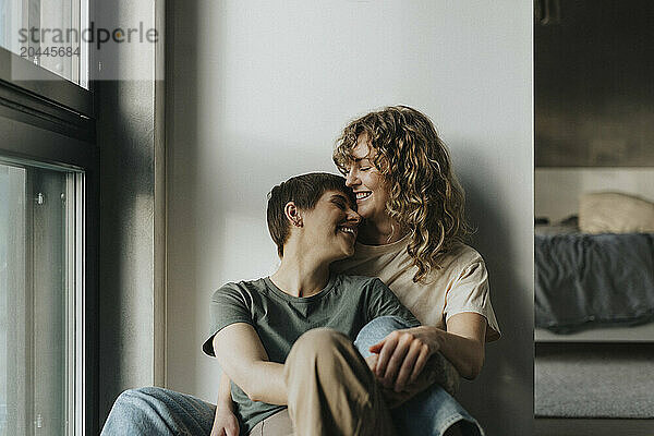 Happy lesbian couple sitting together while leaning on wall at home