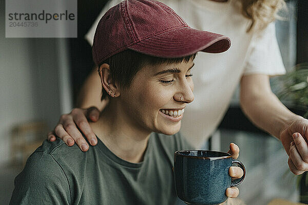 Happy lesbian woman wearing cap holding coffee cup with girlfriend's hand on shoulder