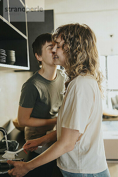 Lesbian woman kissing girlfriend while washing dishes in kitchen at home