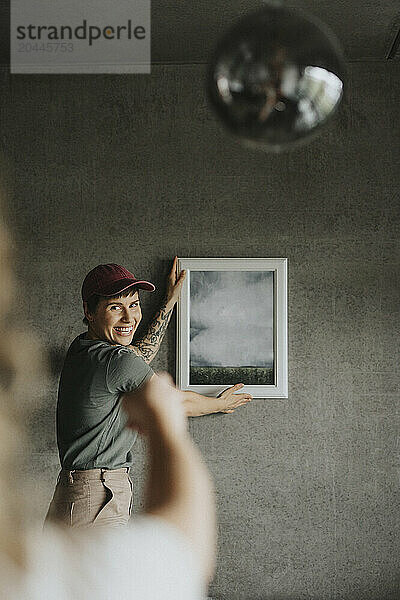 Happy lesbian woman wearing hat taking advice from girlfriend while hanging frame on wall at home