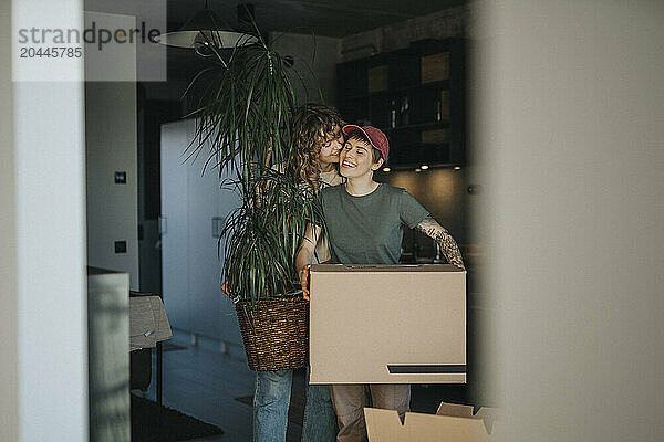 Lesbian woman kissing girlfriend carrying cardboard box during relocation