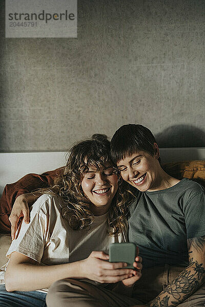 Happy lesbian couple sharing smart phone while sitting at home