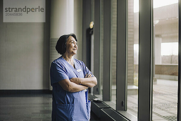 Smiling senior female physician standing with arms crossed near window in hospital