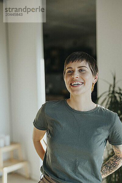 Happy woman with tattooed arm standing with hands behind back at home