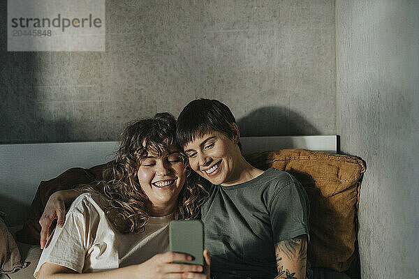 Happy lesbian couple using smart phone while sitting on bed at home