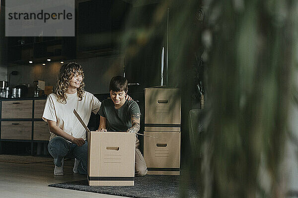 Smiling lesbian woman sitting beside girlfriend packing stuff in cardboard box during relocation