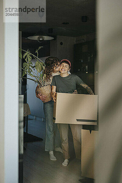 Lesbian woman holding houseplant kissing happy girlfriend carrying cardboard box during relocation