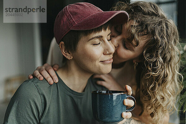 Romantic lesbian woman kissing girlfriend holding coffee cup at home