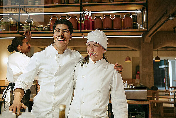 Happy male chef with arm around female chef enjoying while standing in commercial kitchen