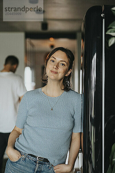 Portrait of beautiful woman leaning on refrigerator with hands in pockets at home