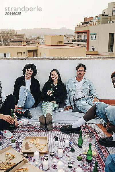 Smiling male and female friends sitting near wall on rooftop during party