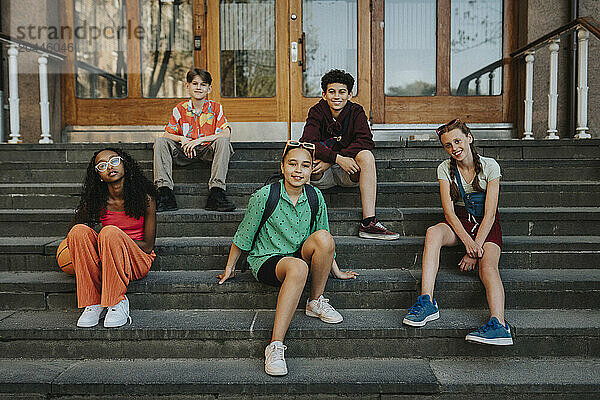 Happy group of friends sitting on staircase of school building entrance