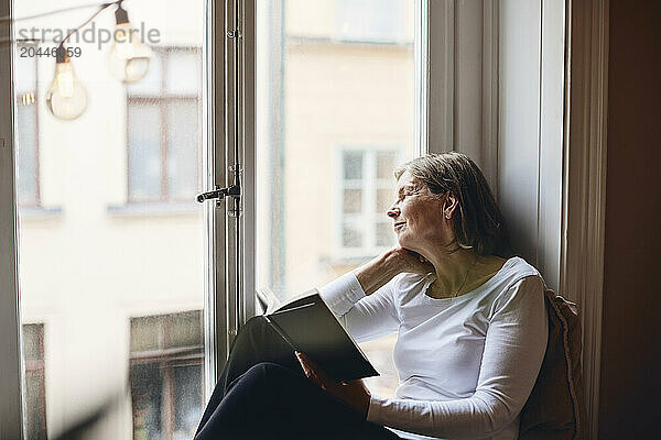 Senior woman looking through window while holding book at home