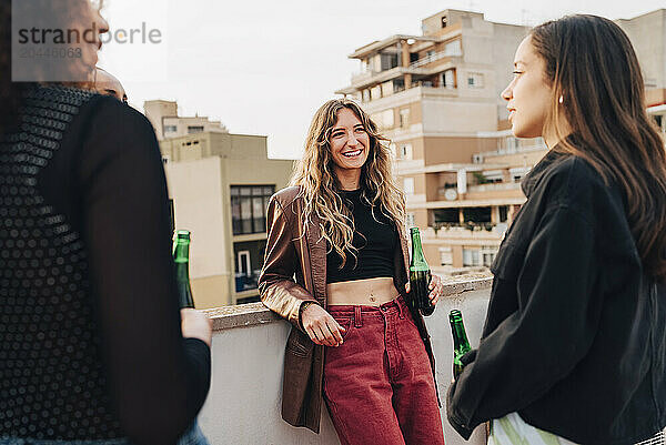 Smiling young woman holding beer bottle and having fun with female friends at rooftop party