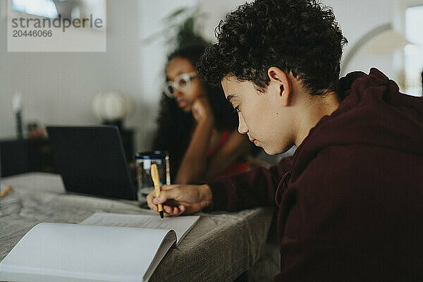 Focused boy with curly hair writing on book while sitting with friend at home