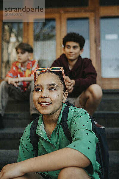 Smiling girl wearing backpack sitting with male friends on staircase in front of school building