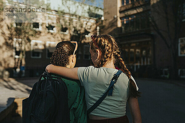 Rear view of girl walking with arm around female friend at school campus