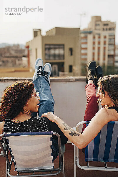 Smiling female friends talking to each other while sitting on chairs at terrace