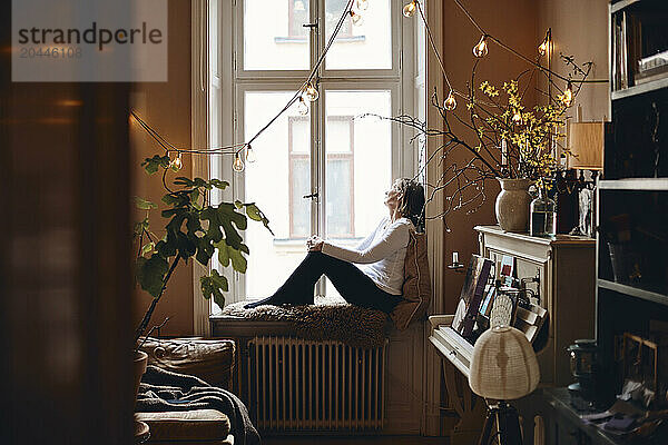 Side view of senior woman sitting on window sill at home