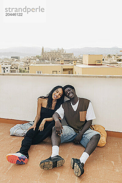 Carefree male and female friends sitting on floor near retaining wall on rooftop