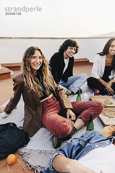 Portrait of smiling woman sitting cross-legged with male and friends during party on rooftop