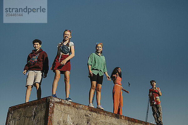 Low angle view of male and female friends standing on wall under clear sky