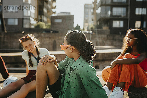 Girl wearing sunglasses looking at friend while sitting on wall at sunny day