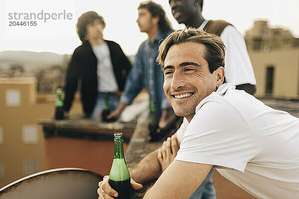 Smiling man holding beer bottle and leaning on railing with friends in background