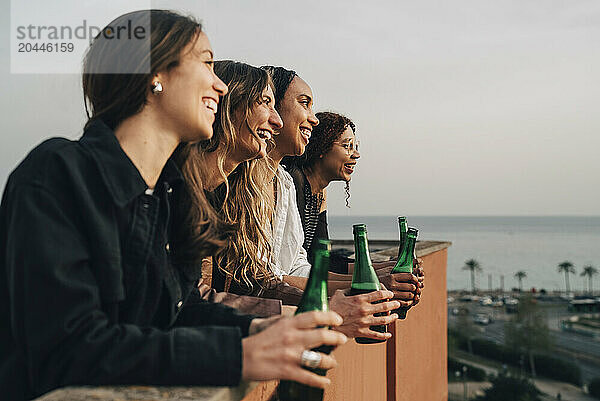 Happy female friends holding beer bottles and leaning on wall of terrace