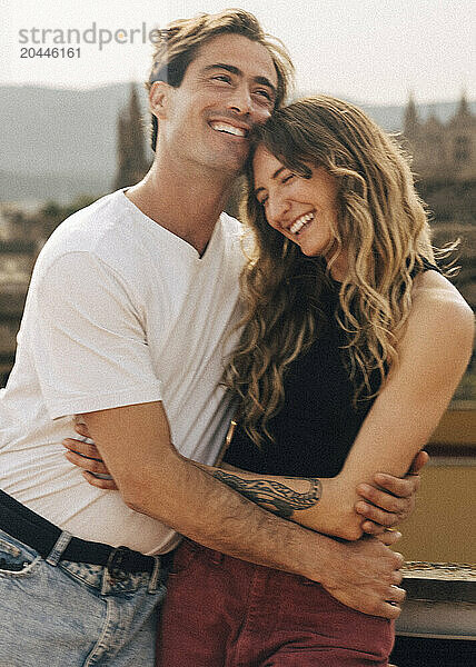 Smiling man embracing happy girlfriend standing on rooftop