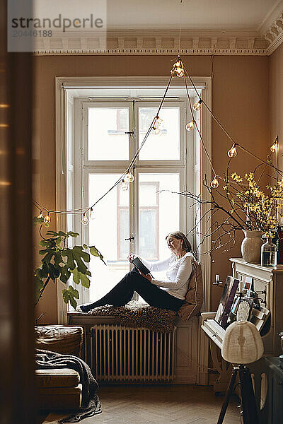 Side view of elderly woman reading book while sitting on window sill