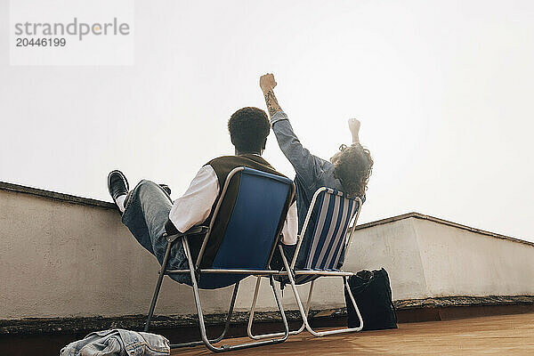 Low angle view of man cheering while sitting with male friend on chair against sky