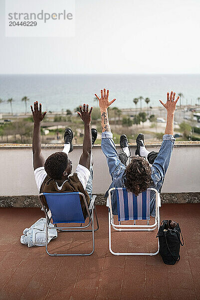 Rear view of carefree male friends sitting with arms raised on chairs at building terrace