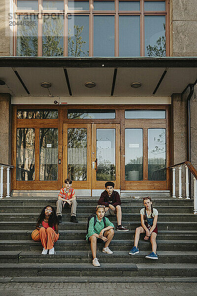 Portrait of male and female friends sitting on staircase in front of school building