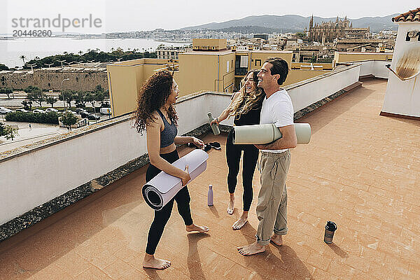 Happy man holding exercise mat and standing with female friends after yoga class on rooftop