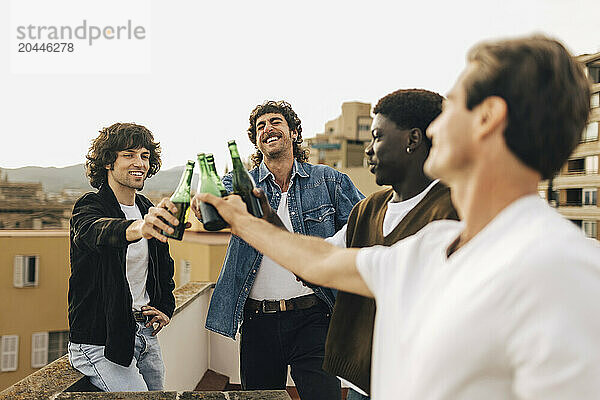 Cheerful male friends toasting beer bottles in party on building rooftop