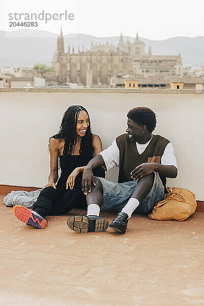 Happy male and female friends sitting near retaining wall on rooftop