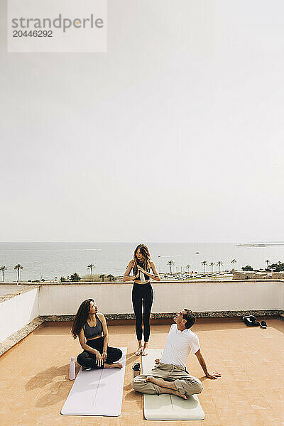 Young woman standing near male and female friends sitting on rooftop during yoga class