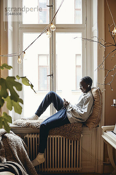 Side view of smiling teenage boy using smart phone while sitting on window sill at home