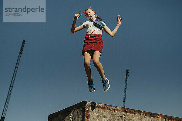 Carefree girl jumping in mid-air while doing peace sign against blue sky