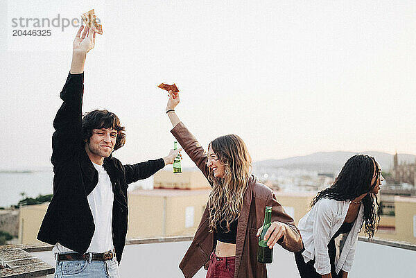 Carefree male and female friends dancing while having snacks at rooftop party