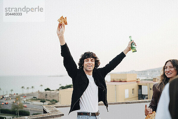 Portrait of carefree man holding beer bottle and pizza slice at rooftop party