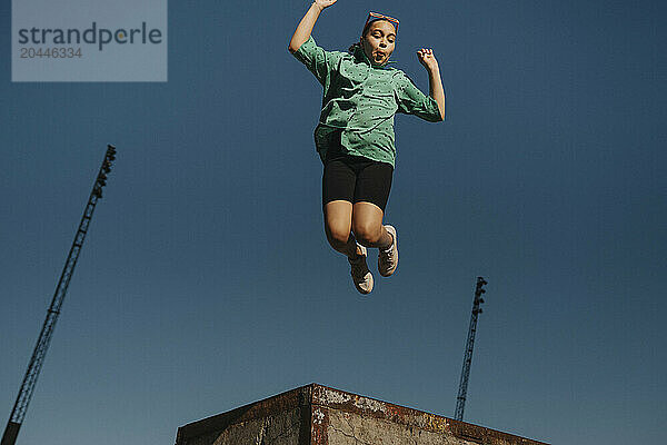 Carefree girl making face while jumping in mid-air against blue sky